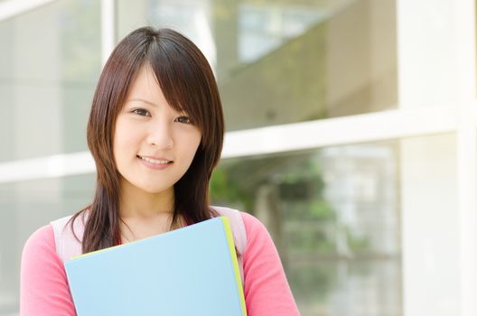 Young Asian adult student standing outside campus building, holding file folder and smiling.