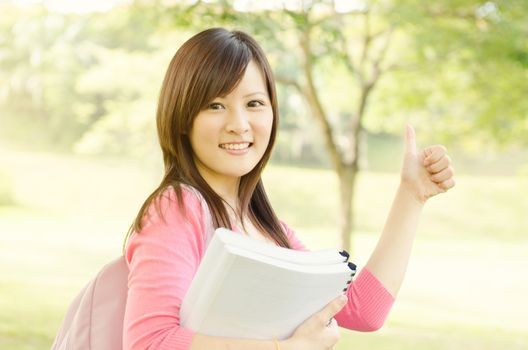 Young Asian college student on campus, holding books and giving a thumb up.