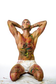 Attractive young man shirtless, skin painted all over with bright Holi colors, looking at camera, sitting, isolated on white background