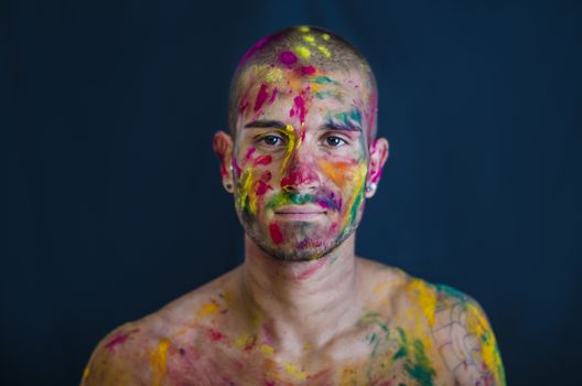 Head and shoulders shot of attractive young man shirtless, skin painted all over with bright Holi colors, looking at camera, isolated on black background