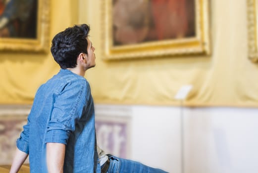 Half Body Shot of a Thoughtful Handsome Young Man, Looking At Painting Sitting on Bench Inside a Museum