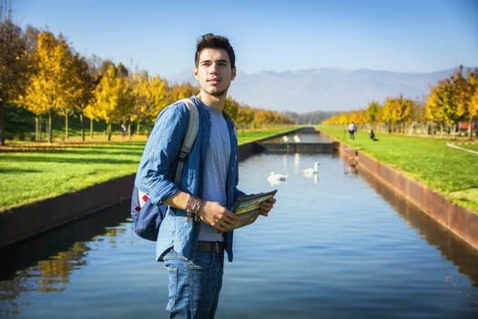 Handsome young man wearing a backpack sightseeing in a lush green autumn park standing alongside a canal with swans, holding a map or brochure looking into the distance as though lost