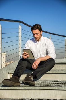 Handsome trendy man wearing white shirt sitting and working, looking down at a tablet computer that he is holding, outdoor on metal stairs step in city setting