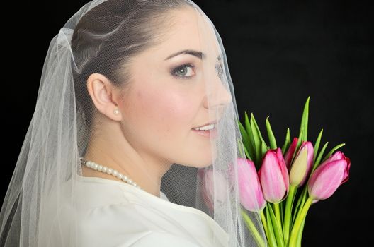 Female model, face covered with veil. Bride holds tulips bouquet. Portrait in studio with black background.
