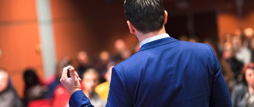 Rear view of speaker giving a talk on corporate Business Conference. Audience at the conference hall. Business and Entrepreneurship event. Panoramic composition.