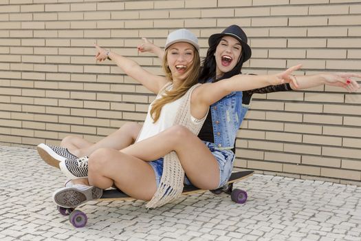 Two beautiful and young girlfriends having fun with a skateboard, in front of a brick wall