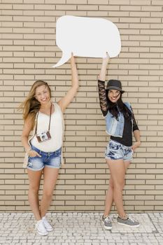 Two beautiful and young girlfriends holding a thought balloon, in front of a brick wall