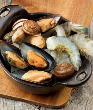 Delicious Raw Seafood with Big Prawns, Squid, Mussels and Snails Ready to Preparing in Black Iron Stewpot closeup on Wooden Cutting Board