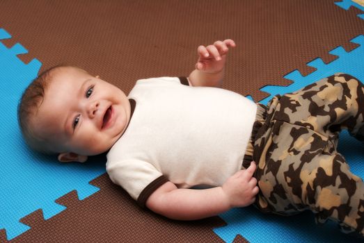 An adorable baby boy of five months lays on his back while giving a happy smile to the camera.