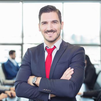 Successful team leader and business owner proudly standing with crossed arms with  coworkers working in office in background. Business and entrepreneurship concept.