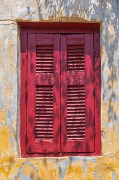 Window with a closed red wooden shutter. Old facade with yellow spots. Shadow of a tree.