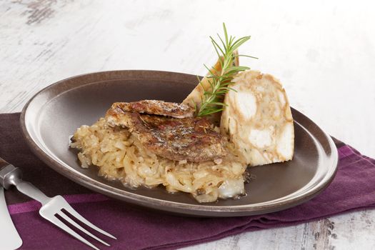 Baked pork chop with dumplings and sauerkraut on plate with silver cutlery on wooden table.