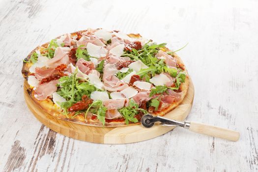 Culinary pizza with prosciutto, dry tomatoes and fresh herbs on wooden cutting board on wooden table. 