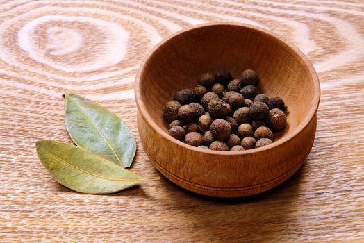 Fragrant black pepper in a wooden bowl and two dried bay leaves on a light wooden table.