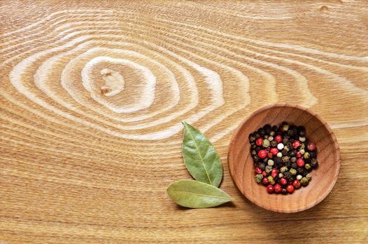 Fragrant colorful pepper in a wooden bowl and two dried bay leaves on a light wooden table. Top view.