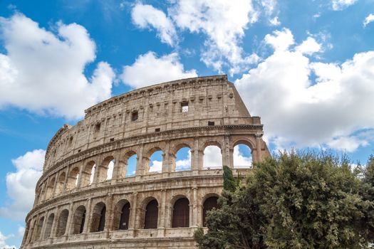 View of ancient amphitheatre of Colosseum built by Vespasian and Titus in Rome, inside trees, on cloudy blue sky background.