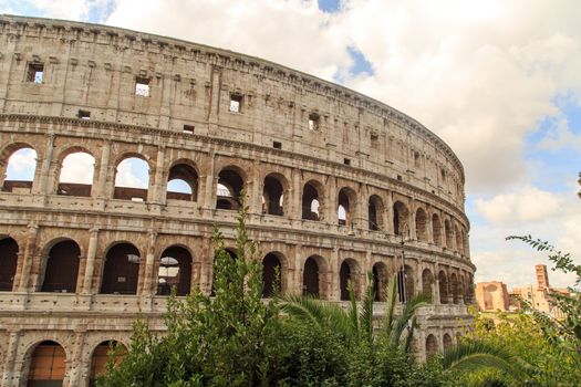 View of amphitheatre of Colosseum built by Vespasian and Titus in Rome, inside trees, on cloudy blue sky background.