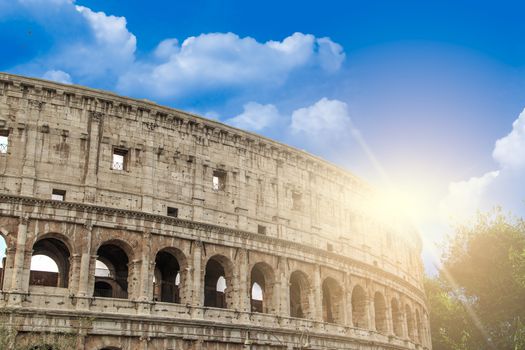 View of amphitheatre of Colosseum built by Vespasian and Titus in Rome, inside trees, on cloudy blue sky with sunshine background.