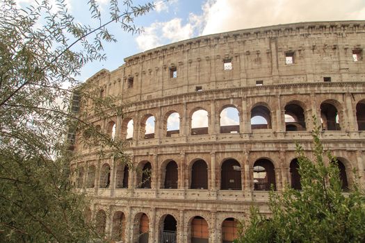 View of amphitheatre of Colosseum built by Vespasian and Titus in Rome, inside trees, on cloudy blue sky background.