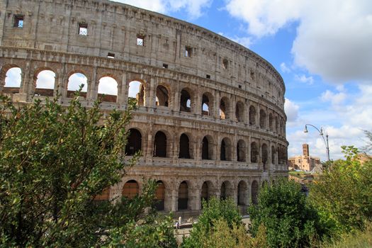 View of amphitheatre of Colosseum built by Vespasian and Titus in Rome, inside trees, on cloudy blue sky background.