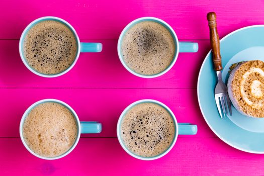 Vibrant overhead image of coffee and cake with four aligned blue cups of frothy black espresso coffee with their handles pointing to chocolate swiss roll cake on a plate on a vivid pink background