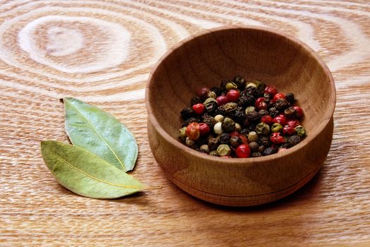 Fragrant colorful pepper in a wooden bowl and two dried bay leaves on a light wooden table.