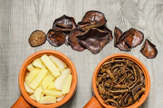 Dried mushrooms and potatoes in a ceramic cups, cut for cooking, and whole mushrooms on old wooden background