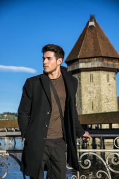 Handsome man standing near metal fence in Lausanne. Famous wooden bridge and Water Tower on background.