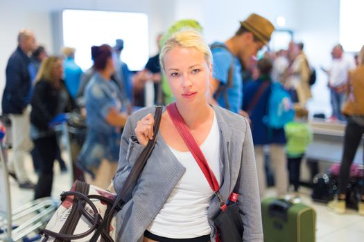 Casually dressed young stylish female traveller waiting in airport arrival terminal hall,  collecting her laggage . Blured background. Can also be used as railway, metro, bus station.