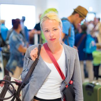 Casually dressed young stylish female traveller waiting in airport arrival terminal hall,  collecting her laggage . Blured background. Can also be used as railway, metro, bus station.