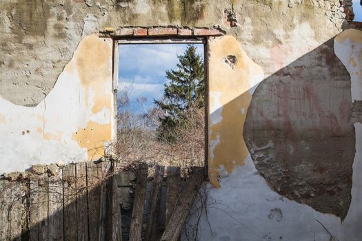 Damaged wall of the room in an abandoned house. Hole instead of the window. Diagonally alighted by sunlight. Nature with a tree and cloudy sky in the window.