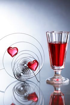 Love concept. Two red glass hearts inside metal springs near wineglass on nice gray background