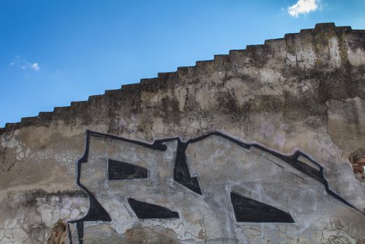 Edge of the roof and a wall with damaged facade and a part of graffiti. Blue sky with small white clouds in the background.