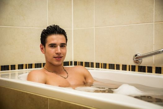 Handsome young man in bathtub at home having bath, washing body and hair with bathfoam and shampoo