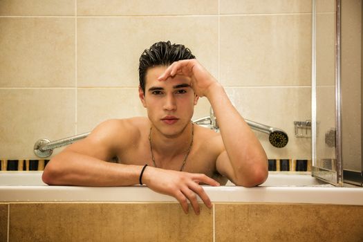 Handsome young man in bathtub at home having bath, washing body and hair with bathfoam and shampoo