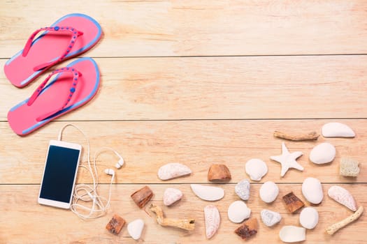 Pictured beach slippers, phone with earphones and shells on wooden boards background.