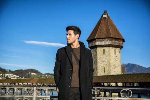 Handsome man standing near metal fence in Lausanne. Famous wooden bridge and Water Tower on background.