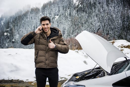 Smiling man talking on cellphone with thumb up near car with open hood. Snowy forest on background