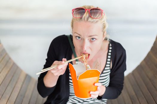 Beautiful young woman, sitting on the bench in park, holding a fast food lunch box, eating noodles from Chinese take-away with traditional wooden chopsticks.