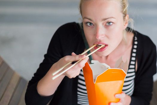 Beautiful young woman, sitting on the bench in park, holding a fast food lunch box, eating noodles from Chinese take-away with traditional wooden chopsticks.