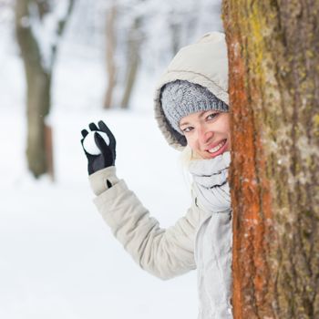 Cute casual young woman hiding behind tree while snowball fighting in winter time.