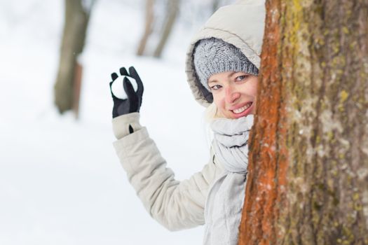 Cute casual young woman hiding behind tree while snowball fighting in winter time.