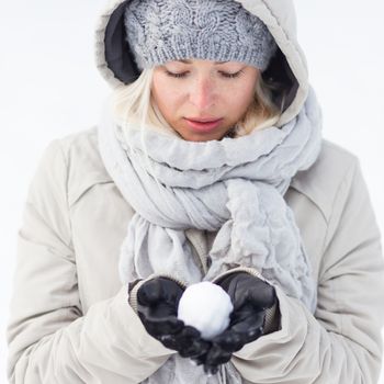 Cute casual young woman wearing glooves, woolen cap and scarf, holding icy snowball in cold winter time. Lady looking down at snowball.