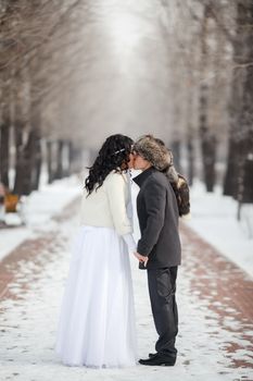 Asian bride and groom kissing in the middle of snowy winter alley. Young man in winter coat and fur hat, bride in white wedding dress with sheepskin. Cold season warmed wedding dresses, wedding coat.