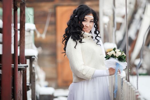 East Asian beautiful brunette bride in the white wedding dress decorated with bandage on her head on a background of buildings in winter and snow. The cold season warmed wedding dresses, wedding coat.