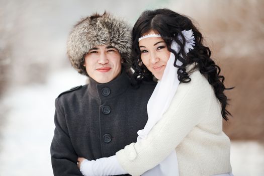 Beautiful wedding couple, asian bride and groom embraced. Young man in winter coat and fur hat, bride in white wedding dress with sheepskin and veil. Cold season warm clothing. Close-up portrait.