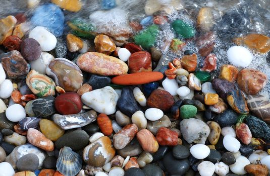 Multicolored sea pebbles and shells in the wave on the shore for background.