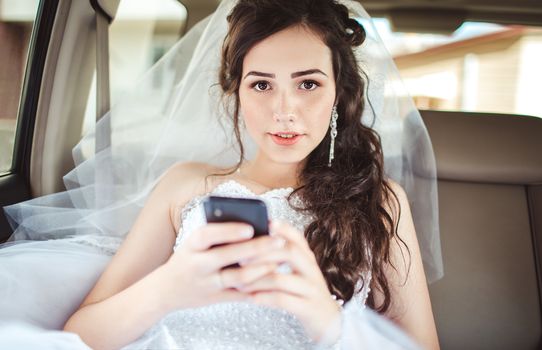 Wedding: beautiful bride sitting in car straight with mobile phone hairstyle and bright makeup. Woman in white dress at wedding day waiting for groom and posing. Concept of love and interest. Newlywed