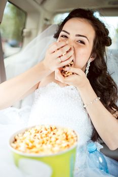 Portrait of beautiful young bride sitting in car straight, greedily eats popcorn before movie, hairstyle and bright makeup. Strong expression, shallow depth of field, focus on eyes and a bit on mouth.