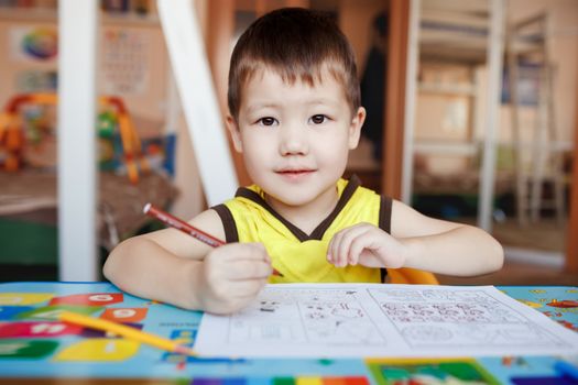 A three year boy in the yellow T-shirt with hood sits at the table, drawing and writing letters with color pencils, looking straight at the camera, interested gaze.